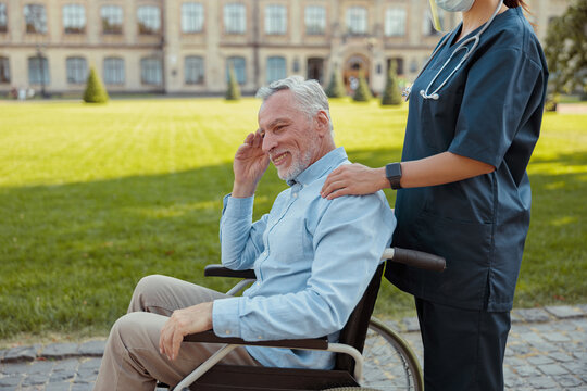 Happy Aged Man, Recovering Patient In A Wheelchair On A Walk With A Nurse Wearing Face Shield And Mask On A Summer Day Outdoors