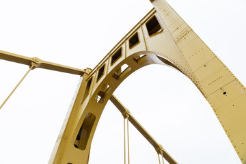 Bright yellow painted upright metal truss of a self anchored suspension bridge against an overcast...