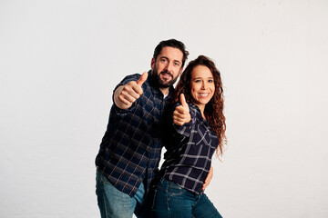 An adult couple in plaid shirts showing thumbs up to the camera and smiling against a white background - 463493032