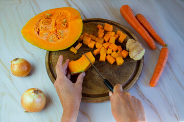 Autumn dishes, pumpkin soup cooking.Female hands are cutting a pumpkin on a wooden plank, onions, ginger and carrots are lying nearby