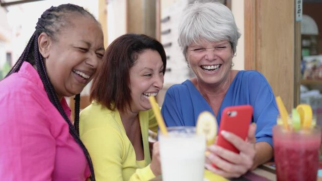Happy multiracial senior having fun using mobile phone at brunch restaurant - Elderly generation people having fun