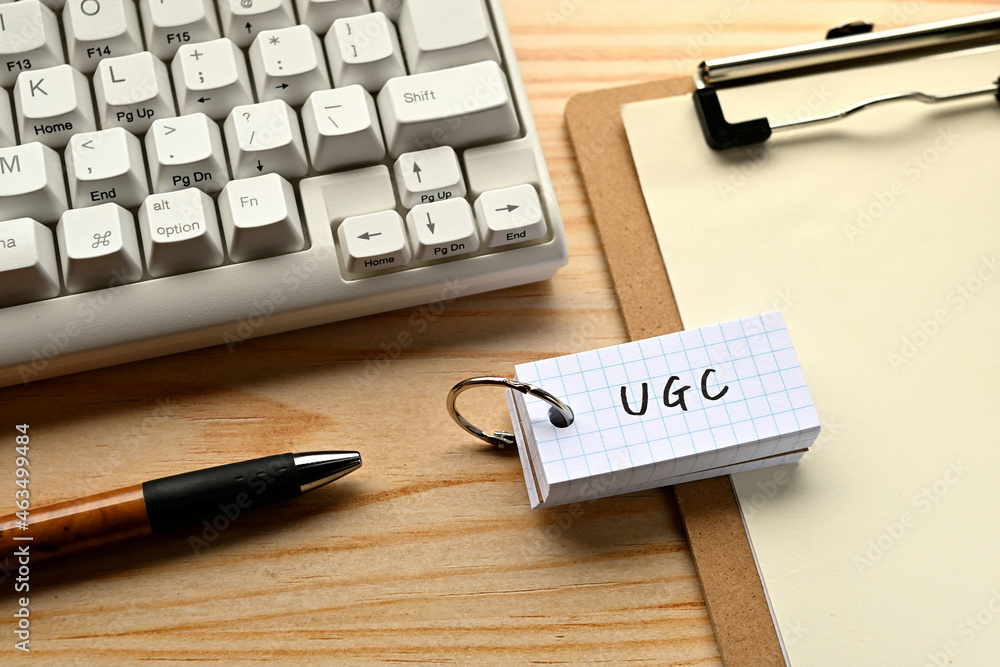 Poster There is a word book with the word of UGC which is an abbreviation for User Generated Contents on the desk with a pen and a keyboard.