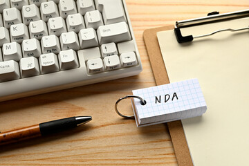 There is a word book with the word of NDA which is an abbreviation for Non-Disclosure Agreement on the desk with a pen and a keyboard.