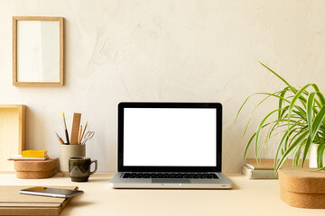 Desk with laptop screen, desk objects, office supplies, books, and plant on a beige clay wall background.	