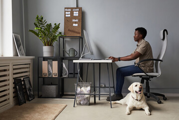 Full length portrait of young African-American man working in office with dog laying on floor, pet...