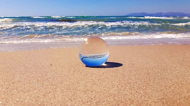 Gentle waves wash around a glass ball in slow motion that lies in the sand in front of the sea.