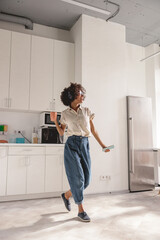 Fototapeta na wymiar Full-length photo of smiling Afro American lady using headphones and dancing on the kitchen while holding smartphone