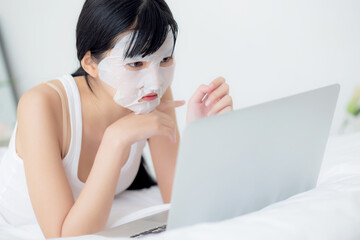 Beautiful young asian woman with sheet facial mask lying working on laptop computer on bed at...