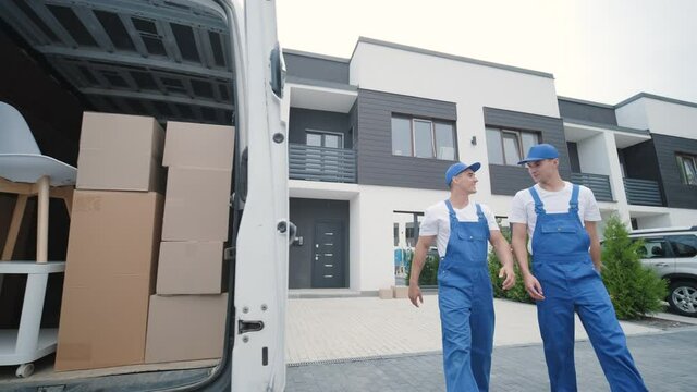 Two young workers of removal company unload boxes and furniture from minibus into customer's home