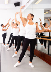 Dancing women engaged in a group class perform the battement tendu movement near a barre, standing in a ballet stance, where ..the choreographer helps to perform the exercise correctly
