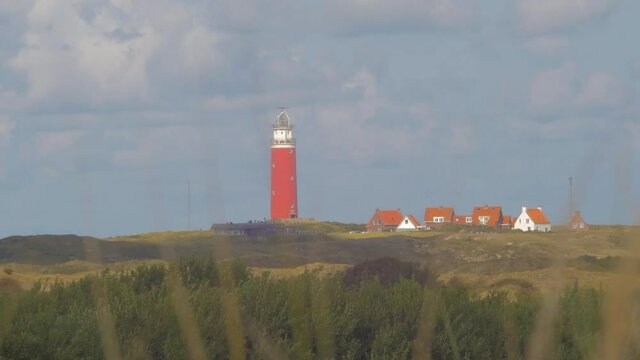 Steady Tele shot of Waving Marram grass in the foreground. In the distance we see a lighthouse in the dunes together with some builings. The sky is blue with beautiful clouds passing by.