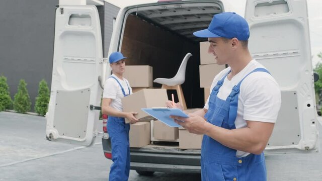 Two young workers of removal company unload boxes and furniture from minibus into customer's home