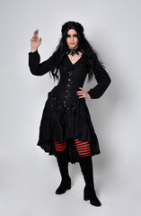 Full length portrait of dark haired woman wearing  black victorian witch costume with corset,  standing pose with  gestural hand movements,  against studio background.