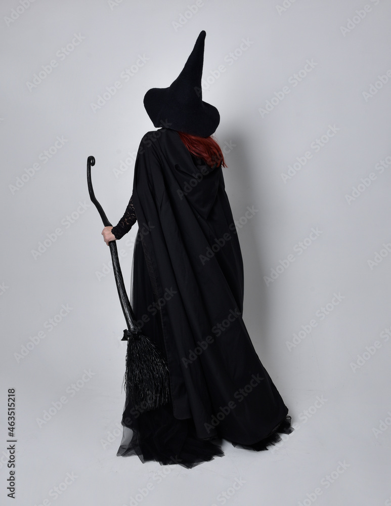 Canvas Prints Full length portrait of dark haired woman wearing  black victorian witch costume with  cloak and pointy hat.  standing pose, back view,  with  gestural hand movements,  against studio background.