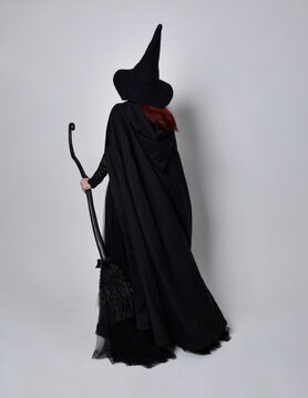 Full length portrait of dark haired woman wearing  black victorian witch costume with  cloak and pointy hat.  standing pose, back view,  with  gestural hand movements,  against studio background.