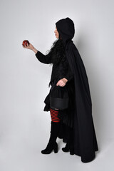 Full length portrait of dark haired woman wearing  black victorian witch costume with  cloak and pointy hat.  standing pose, back view,  with  gestural hand movements,  against studio background.