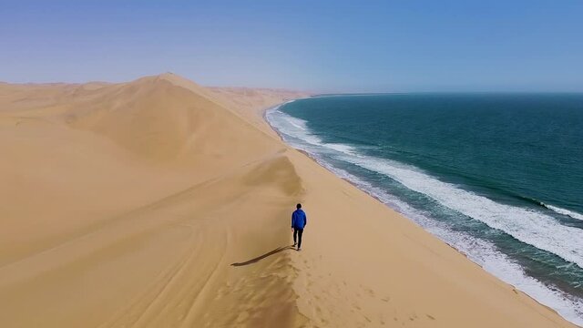 Drone flies over man walking on crest of dune next to ocean, barren landscape
