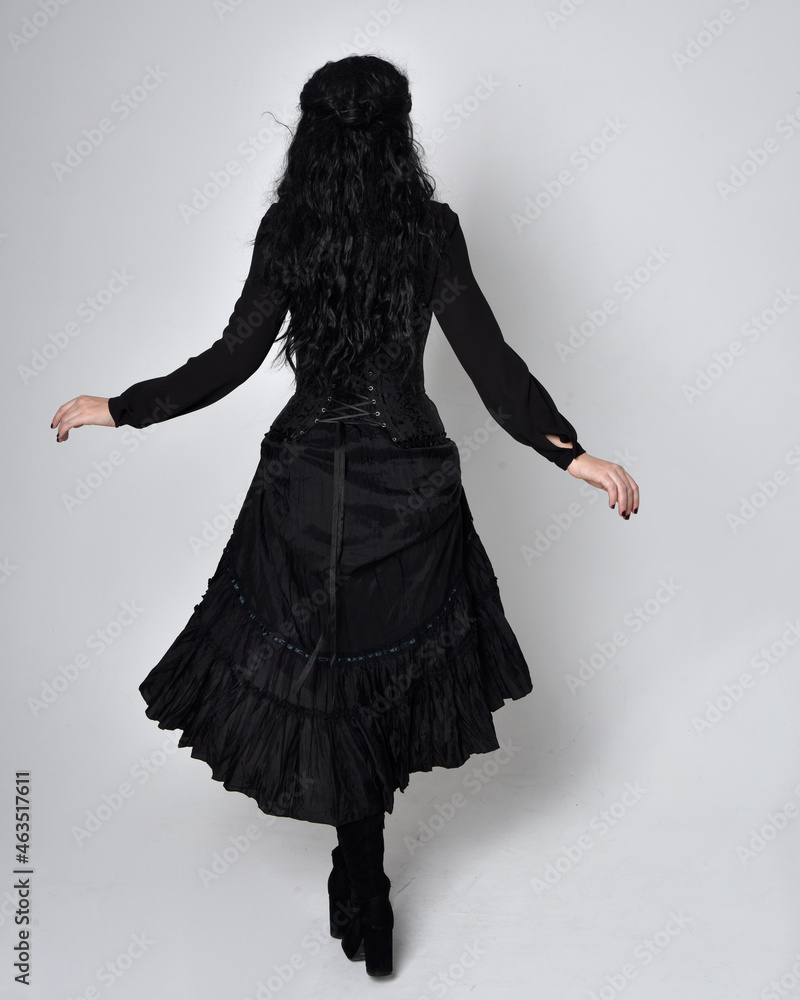 Canvas Prints Full length portrait of dark haired woman wearing  black victorian witch costume.  standing pose, back view,  with  gestural hand movements,  against studio background.