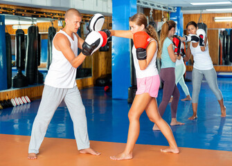Determined young slim woman in boxing gloves learning to punch, exercising on focus mitts held by...