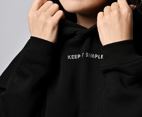 Closeup of chest and neck of woman in trendy black hoodie with inscription Keep it simple, holding her cowl with hands, putting it on or taking off