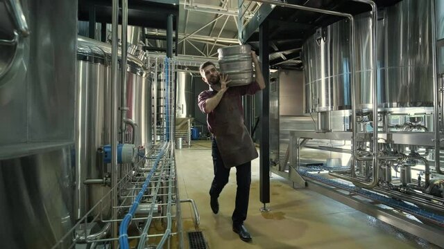 Young brewer wearing a leather apron carries a barrel of craft beer at a modern brewery