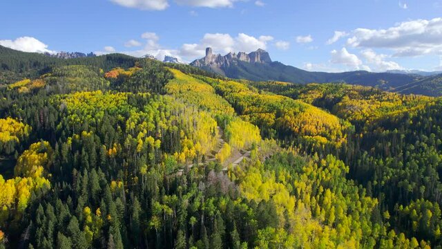 Autumn landscape in Colorado with yellow aspens and jagged mountain peaks in distance. Aerial 4K drone video with two cars driving along dirt road near hairpin turn.