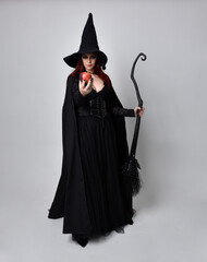 Full length portrait of dark haired woman wearing  black victorian witch costume.  standing pose, holding a broomstick  with  gestural hand movements,  against studio background. - obrazy, fototapety, plakaty