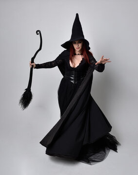 Full length portrait of dark haired woman wearing  black victorian witch costume.  standing pose, holding a broomstick  with  gestural hand movements,  against studio background.