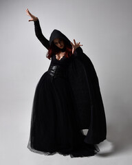 Full length portrait of dark haired woman wearing  black victorian witch costume with a flowing  cloak.  standing pose, with  gestural hand movements,  against studio background.