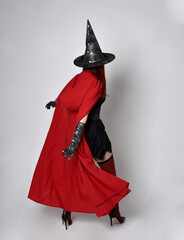 Obraz premium Full length portrait of dark haired woman wearing black victorian witch costume with a flowing cloak. standing pose, with gestural hand movements, against studio background.