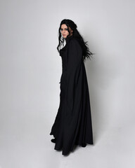 Naklejka premium Full length portrait of dark haired woman wearing black victorian witch costume with a flowing cloak. standing pose, with gestural hand movements, against studio background.