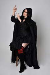 Full length portrait of dark haired woman wearing  black victorian witch costume with a flowing  cloak.  standing pose, with  gestural hand movements,  against studio background.