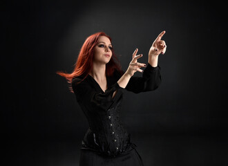 close up portrait of red haired woman wearing  black victorian witch costume.  standing pose, with  gestural hand movements,  against dark studio background.