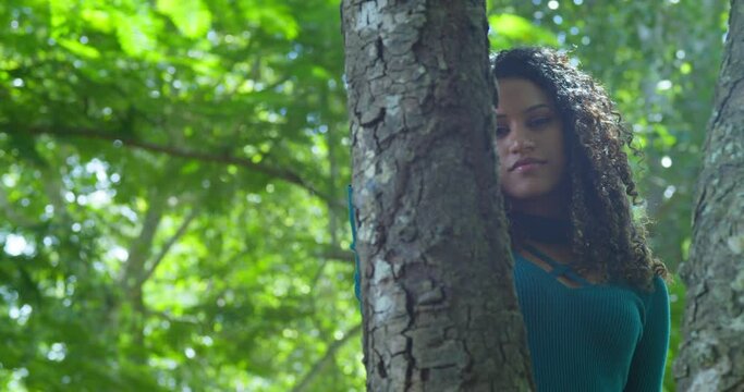 A young curly hair girl with the sun light shine on her hair and face while standing behind a tree trunk