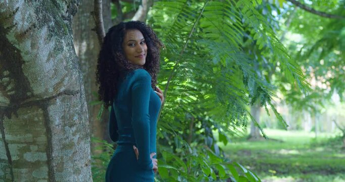 A slim sexy curly hair model stands near a large tree trunk on the tropical island of Trinidad