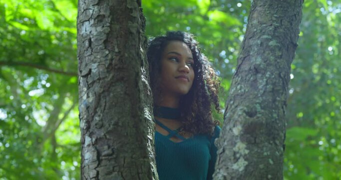 A young sexy girl looks into the sun rays while standing behind a tree trunk