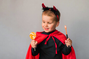 Child with horns on his head and with candy and a brush in his hands is isolated on a grey background on Halloween. Health, children's hygiene, holidays concept.