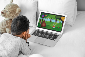 Little African-American boy with laptop watching cartoons on bed