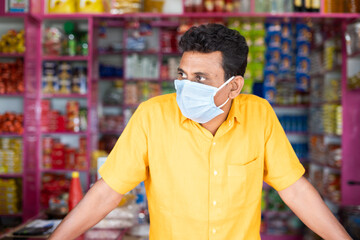Merchant with mdical face mask at groceries or Kirana store waiting for customers by looking around...