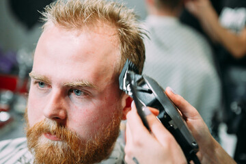 a red-haired adult man gets a haircut with a machine in a barbershop
