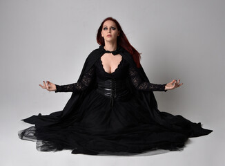 Full length portrait of dark haired woman wearing  black victorian witch costume  sitting pose, with  gestural hand movements,  against studio background.