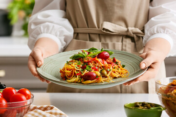Woman holding plate with tasty Pasta Puttanesca at table in kitchen, closeup
