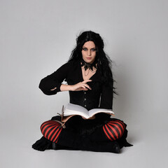 Full length portrait of dark haired woman wearing  black victorian witch costume  sitting pose,...