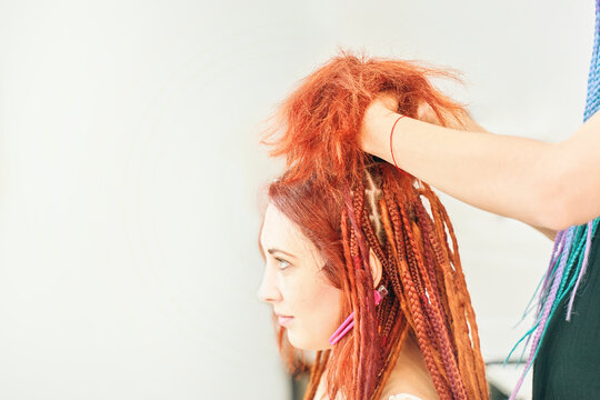 Process of braiding braids on head in beauty salon close up. Red-haired girl makes braids-dreadlocks. Dreadlocks from kanekalon. Cool fashion hairstyle made of artificial hair. Barber services.