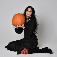 Full length portrait of dark haired woman wearing  black victorian witch costume  sitting pose wit...