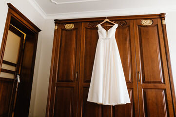 Fashionable beautiful classic lace silk wedding dress hanging on hanger in hotel wooden room. morning preparation wedding concept. vintage wedding gown.