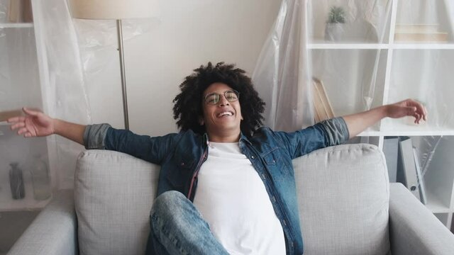 New home. Happy relocation. Real estate. Satisfied relaxed Arab guy hopping falling on cozy couch in light place living room interior with polyethylene isolated furniture.