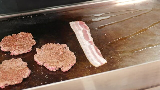 Burger patties lie on a grill. The cook adds bacon and lets it brown.