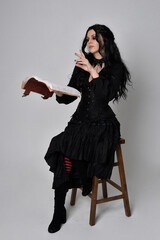 Full length portrait of dark haired woman wearing  black victorian witch costume.  sitting pose on...