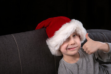 Boy in Santa hat is showing good, thumbs up sitting on the sofa on black background